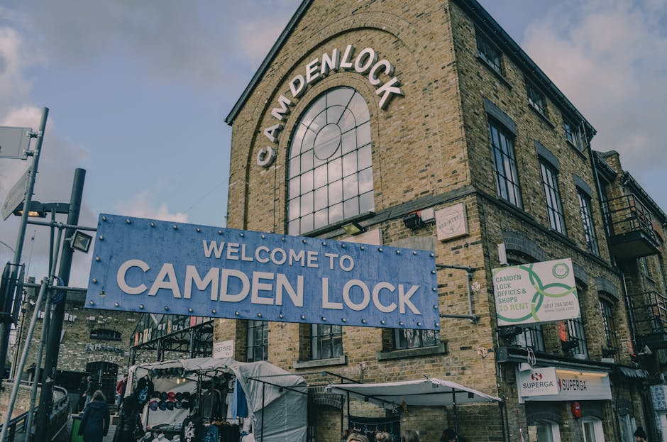 A multi-story brick building with large, arched windows featuring black frames and sills, situated at a street corner in Camden Lock. Prominently displayed on the front of the building is a large blue sign with white and light blue lettering that reads 'WELCOME TO CAMDEN LOCK', supported by metal brackets. Above it, an arched window with a grid pattern reflects a cloudy sky, adding an atmospheric element to the scene. To the right of the main sign, a smaller rectangular sign advertises 'CAMDEN LOCK OFFICES & SHOPS TO RENT', with a green logo and contact information. At the street level, a white canopy covers a vendor stall with a variety of merchandise, including hats and clothing items, which are partially visible. The surrounding environment includes street lamps and metal railings, suggesting an active marketplace or commercial area. The lighting is natural, with diffused sunlight casting soft shadows, creating a lively yet organized ambiance typical of a popular visitor and local destination known for independent shops and outdoor stalls, aligning with the context of private waste handling and rubbish removal in the area.