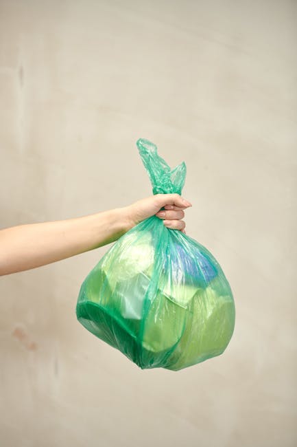 A human hand is holding a green plastic rubbish bag by its twisted knot, with the bag appearing full and slightly translucent, revealing some contents inside. The background is a plain, light-colored wall with a neutral tone and no visible environment or setting. The bag is made of thin, flexible plastic with creases and folds, and the hand holds it firmly from the top, indicating an intent to dispose of household waste or general rubbish. This scene exemplifies an informal task of rubbish disposal, related to private waste handling or non-utility collection, which aligns with services like those offered by Rubbish Clearance Camden in managing domestic or flat waste. The lighting is soft and diffuse, emphasizing the textures of the plastic and the natural colour of the waste bag. The image avoids any promotional details and stays focused on the act of rubbish disposal, supporting the context of independent collection or waste removal services suitable for flat or residential environments.