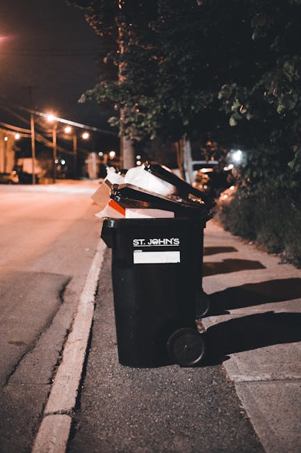 A large black wheelie bin with the label 'ST. JOHN'S' placed on a street curb at night, filled with various rubbish including cardboard boxes, plastic bags, and paper. The bin sits on a concrete sidewalk next to a white painted line marking the edge of the road. In the background, there are streetlights casting warm orange light, illuminating tree foliage and parked vehicles along the quiet street. The scene suggests a late evening or night-time setting, with shadows cast by the bin and surrounding objects, and an environment typical of urban or suburban areas where private waste disposal services might be employed for rubbish collection. The bin and its contents visually represent the accumulation of household or commercial waste awaiting collection, relevant to independent waste clearance or rubbish removal services such as those provided by Rubbish Clearance Camden.