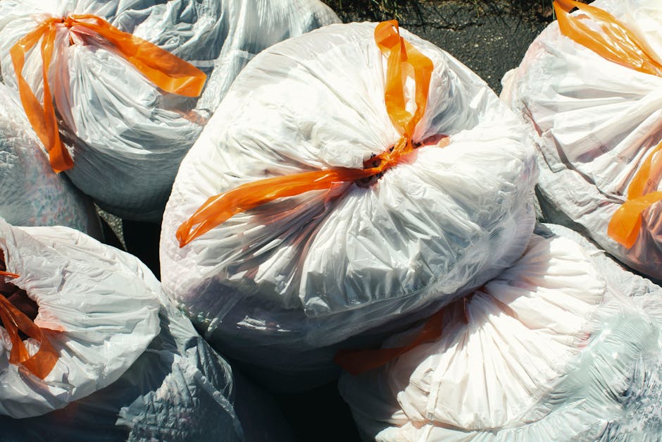 A human hand is holding a green plastic rubbish bag by its twisted knot, with the bag appearing full and slightly translucent, revealing some contents inside. The background is a plain, light-colored wall with a neutral tone and no visible environment or setting. The bag is made of thin, flexible plastic with creases and folds, and the hand holds it firmly from the top, indicating an intent to dispose of household waste or general rubbish. This scene exemplifies an informal task of rubbish disposal, related to private waste handling or non-utility collection, which aligns with services like those offered by Rubbish Clearance Camden in managing domestic or flat waste. The lighting is soft and diffuse, emphasizing the textures of the plastic and the natural colour of the waste bag. The image avoids any promotional details and stays focused on the act of rubbish disposal, supporting the context of independent collection or waste removal services suitable for flat or residential environments.
