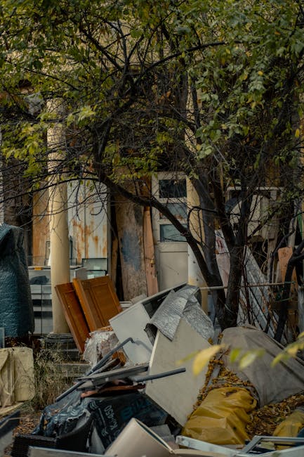 The image shows an outdoor scene with a pile of discarded household items and rubbish situated beneath a tree with leafy branches. In the foreground, there are various materials including a white appliance that appears to be broken or discarded, a yellow bag, black plastic wrap, and fragmented wooden panels stained in shades of orange-brown. Some pieces are leaning against each other and partially obscured by the surrounding environment. The background features a building with weathered, peeling paint in cream and rust tones and a rusty metal fence, suggesting an area used for waste accumulation or clearance. The scene is lit with natural daylight, and the overall setting resembles a private property or alleyway where rubbish has been temporarily gathered, possibly as part of a local clearance or independent disposal process supported by companies such as Rubbish Clearance Camden. The clutter and placement of items visually emphasize the context of waste removal services aimed at clearing outdoor spaces.