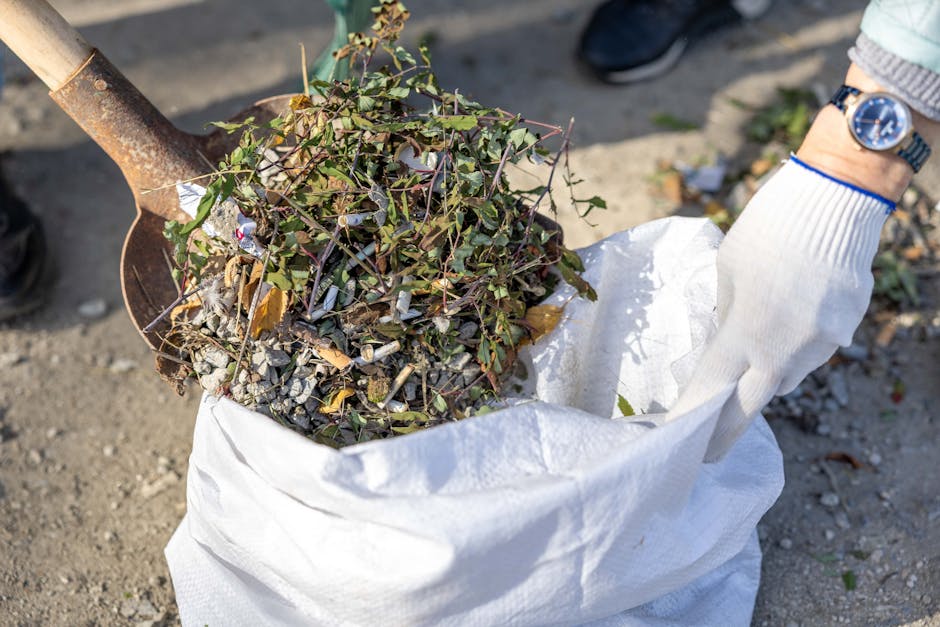 A person wearing a white glove is holding a large white sack filled with miscellaneous garden waste, including small leafy branches, twigs, and gravelly soil. The sack's fabric appears durable and slightly crinkled, with the waste visible at the top. Adjacent is a rusted metal shovel partially inserted into the sack, with a wooden handle extending out of the frame. The scene is outdoors on a gravel driveway or pathway, with some scattered small stones and bits of plant debris on the ground. The individual’s wristwatch and a light-colored sleeve of clothing are visible on the right side of the image. The background is blurred but suggests an open environment suitable for garden clearance or waste collection, which relates to private rubbish removal services like those offered by Rubbish Clearance Camden for garden and general waste disposal, emphasizing the context of independent waste handling or on-site clearance tasks.
